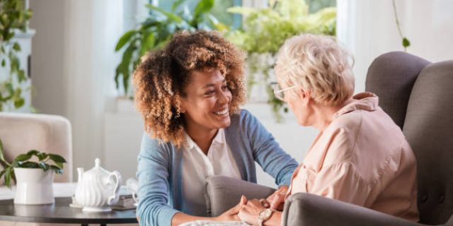 Female home caregiver talking with senior woman, sitting in living room and listening to her carefully.